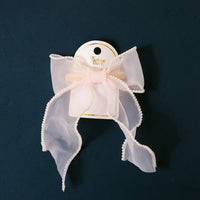 The Blush Sheer Pearl Hair Bow by the-darling-effect features a light pink, sheer ribbon with pearl trim, attached to a white and gold tag—an elegant addition to your collection of bow clips.