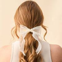 A person with long, wavy, light brown hair is shown from behind wearing the-darling-effect's Cream Sheer Pearl Hair Bow, a delicate clip with decorative edges. They pair it with a sleeveless white top, highlighting this elegant accessory.