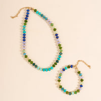 A gold chain necklace and playful jewelry, including The Darling Effect's True Blues Stone Candy Bracelet with colorful blue, green, and purple beads, are laid out against a cream background.