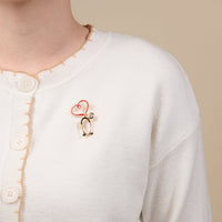 The Darling Effect’s Nurse Love Timeless Brooch—a crystal-accented, gold and red stethoscope brooch with a heart—adorns a white cardigan.