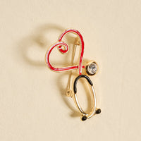 The Darling Effect’s Nurse Love Timeless Brooch features a gold stethoscope design with a red heart outline and clear gemstone, shown on a light beige background—an elegant keepsake accessory.