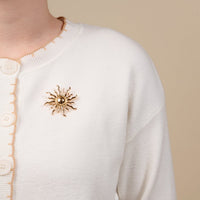 The Darling Effect’s Sunshine Timeless Brooch is pinned to a cream cardigan with beige trim and buttons, bringing vintage-inspired style to the look.