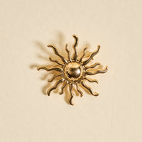 The Darling Effect's Sunshine Timeless Brooch is a gold, sun-shaped vintage-inspired accessory with wavy rays and a round center, beautifully showcased against a light beige background.