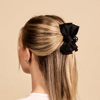 A woman with straight blonde hair, styled in a half-up twist and secured with The Darling Effect's Black Mini Satin Bow Claw Clip, is shown from the back against a beige background. She wears small hoop earrings and a white top.