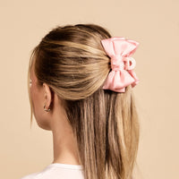 A person with blonde hair wears The Darling Effect's Blush Mini Satin Bow Claw Clip—a large, pale pink bow with a satin finish and a blush claw backing.