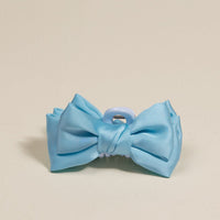 The Darling Effect’s Blue Mini Satin Bow Claw Clip features a light blue satin bow on a mini clip, shown against a beige background. A pretty addition to your hair accessories collection.