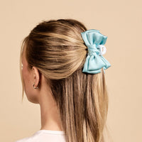 A woman with straight blonde hair, seen from behind, wears The Darling Effect’s Blue Mini Satin Bow Claw Clip and two small hoop earrings in her left ear, elegantly displaying the hair accessory against a beige background.