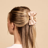 A person with long, straight, blonde hair styled in a half-updo, secured with The Darling Effect’s Cheetah Mini Satin Bow Claw Clip. The background is a solid, neutral beige.