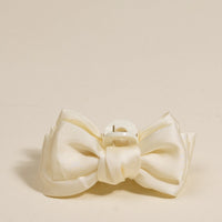 The Darling Effect’s Cream Mini Satin Bow Claw Clip features a cream-colored satin bow and sturdy plastic clasp, shown against a plain beige background.