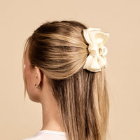 A woman with straight blonde hair styled in a half-updo is facing away from the camera, wearing The Darling Effect's Cream Mini Satin Bow Claw Clip, shown against a beige background.