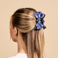 A woman with straight blonde hair styled half-up, half-down secures it with The Darling Effect’s Endless Daydream Navy Mini Satin Bow Claw Clip, which features a blue and white floral pattern. She is seen from behind against a beige background.