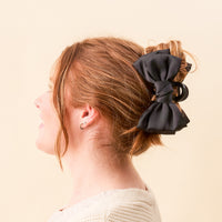 Side profile of a woman with auburn hair in a messy bun, secured by The Darling Effect's Black Satin Bow Claw Clip. She completes her look with a cream knit sweater and small hoop earrings.