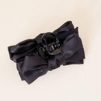 The Darling Effect Black Satin Bow Claw Clip features a large, elegant satin bow—perfect for stylish updos. Shown on a light beige background.