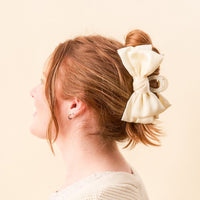 A woman with red hair wears The Darling Effect Cream Satin Bow Claw Clip against a light peach background.