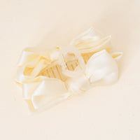 The Darling Effect’s Cream Satin Bow Claw Clip features two large satin-finish bows and is ideal for updos, adding a charming, elegant touch to any hairstyle. Displayed on a light, textured surface.