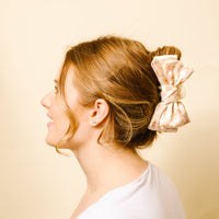A woman with light brown hair in a bun, secured with the-darling-effect's Endless Daydream Cream Satin Bow Claw Clip. She smiles in a white top, viewed from the side against a light beige background.