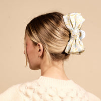 A woman's blonde hair is styled in a bun and secured with The Darling Effect's Poppin' Petals Aqua Satin Bow Claw Clip. She wears a cream-colored sweater.