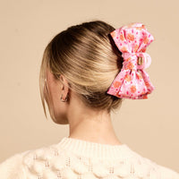 A woman with blonde hair in a low bun, secured by The Darling Effect's Poppin' Petals Hot Pink Satin Bow Claw Clip, wears a cream sweater and small hoop earrings against a beige background.