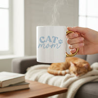 A hand holds The Darling Effect’s Cat Mom 11 oz Cozy Cute Ceramic Mug, featuring blue text and a paw print with a gold handle, as steam rises. In the background, an orange cat naps on a blanket-covered sofa—ideal for coffee or tea moments.