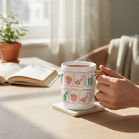 A hand holds The Darling Effect’s Mahjong Tiles 11 oz Cozy Cute Ceramic Mug, featuring colorful tile designs and a gold handle, resting on a coaster beside an open book, potted plant, and soft fabric on a sunlit wooden table.