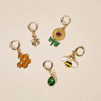 Five gold hoop charms from The Darling Effect’s Busy Bee The Ultimate Charm Pack on a beige background, featuring a flower, sunflower, honeycomb, green gemstone, and a bee with white wings and yellow stripes.