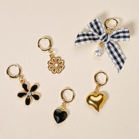 Five gold huggie charm earrings—black enamel daisy, gold flower, black crystal heart, gold puffy heart, and black-and-white gingham bow with crystal drop—by The Darling Effect’s Midnight Moments The Ultimate Charm Pack. Mix and match with your favorites!.