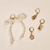 Four gold-tone earrings from The Darling Effect’s Pearl Party The Ultimate Charm Pack are displayed on a cream background, featuring a pearl beaded bow, yellow teardrop gem, faceted square gem, and golden flower.