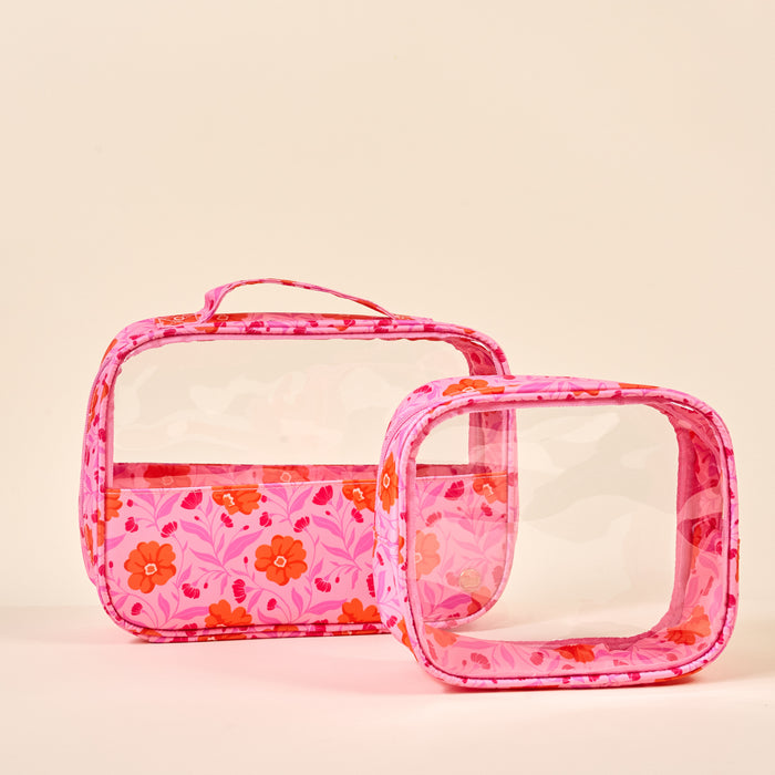 The Darling Effect's Poppin' Petals Hot Pink Clearly Chic Cosmetic Bag Set features two transparent rectangular toiletry bags with pink floral trim and handles, one upright and one on its side, both against a beige background.