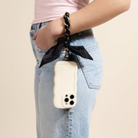 A person wears light blue jeans and a pale pink shirt, holding a smartphone with The Darling Effect's Black Call Me Later Phone Scarf—a wavy-edged white case with a gold-tone chain strap and black ribbon—chicly hanging from their wrist.