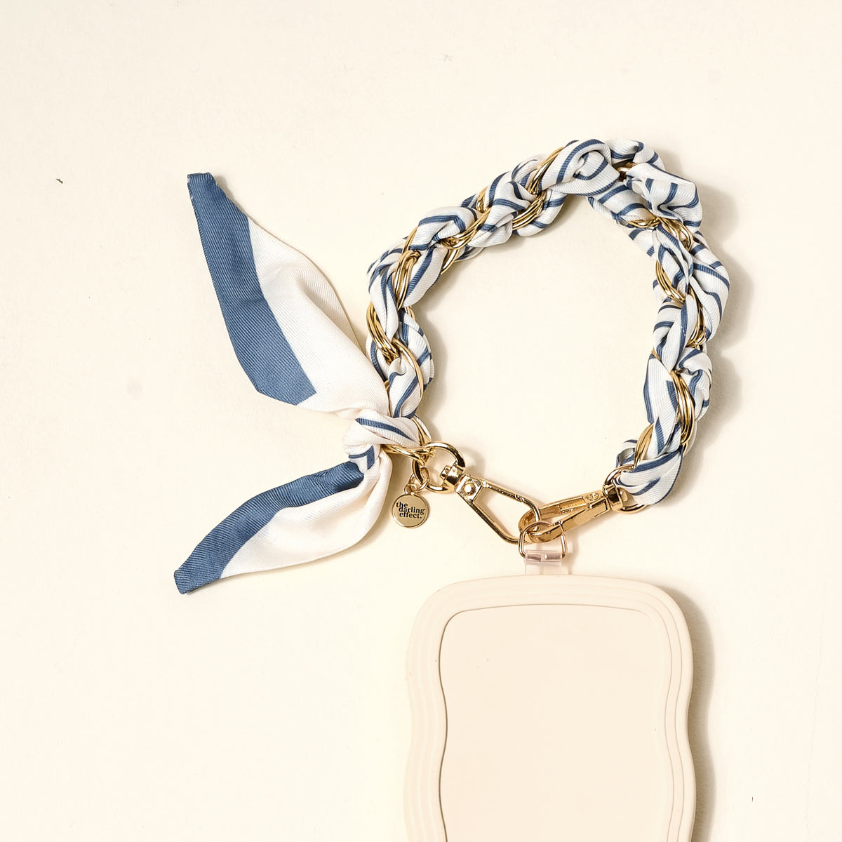 The Darling Effect’s Bluebell Stripe Call Me Later Phone Scarf features a braided blue, white, and gold ribbon with gold-tone chain hardware, a heart charm, and a cream wavy-edged tag, displayed on a light background.