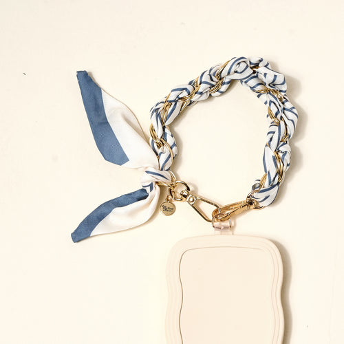The Darling Effect’s Bluebell Stripe Call Me Later Phone Scarf features a braided blue, white, and gold ribbon with gold-tone chain hardware, a heart charm, and a cream wavy-edged tag, displayed on a light background.