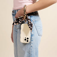 A person in light blue jeans and a light pink shirt holds a cream phone with a wavy case, attached to a gold-tone chain strap featuring The Darling Effect's Cheetah Call Me Later Phone Scarf.