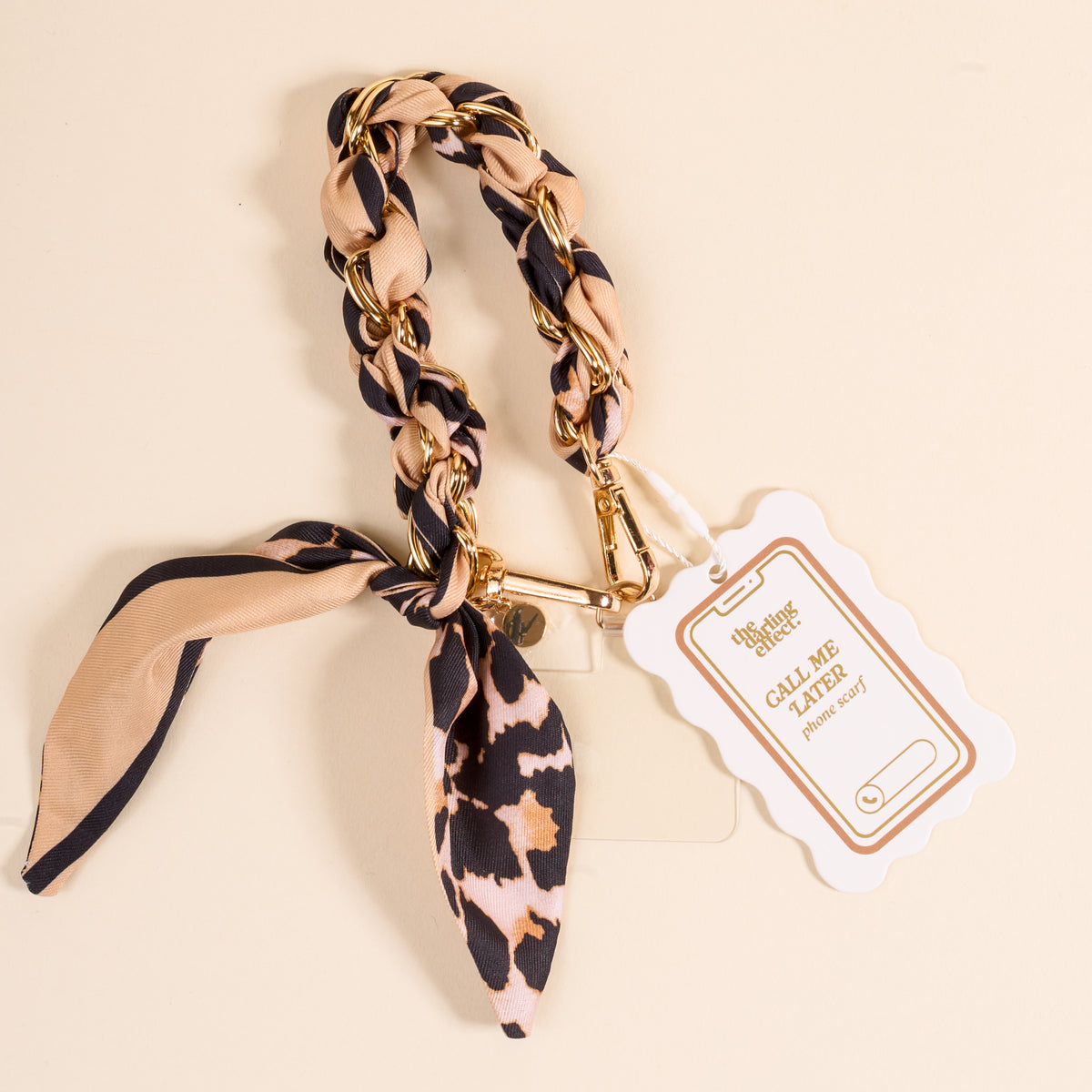 The Darling Effect’s Cheetah Call Me Later Phone Scarf features a gold-tone chain with beige and black cheetah-print fabric tied in a bow, plus a “CALL ME LATER Phonegrip” tag—the perfect stylish accessory for your device.