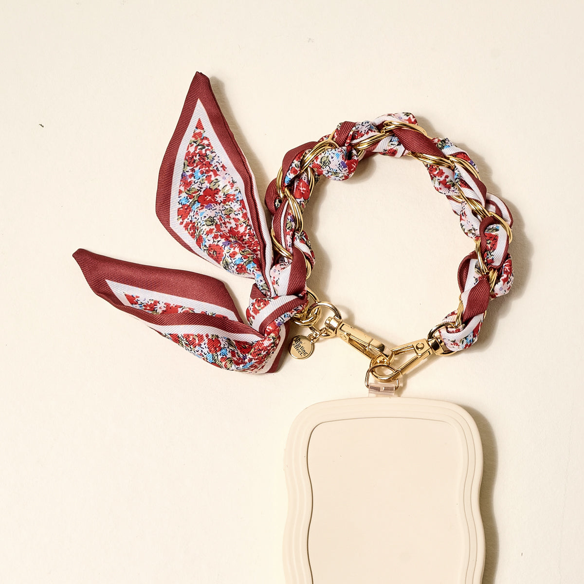 The Crimson Rose Call Me Later Phone Scarf by The Darling Effect, featuring a beige rectangular wavy-edged keychain and gold-tone chain bracelet, is styled on a light cream background—a chic accessory for any look.