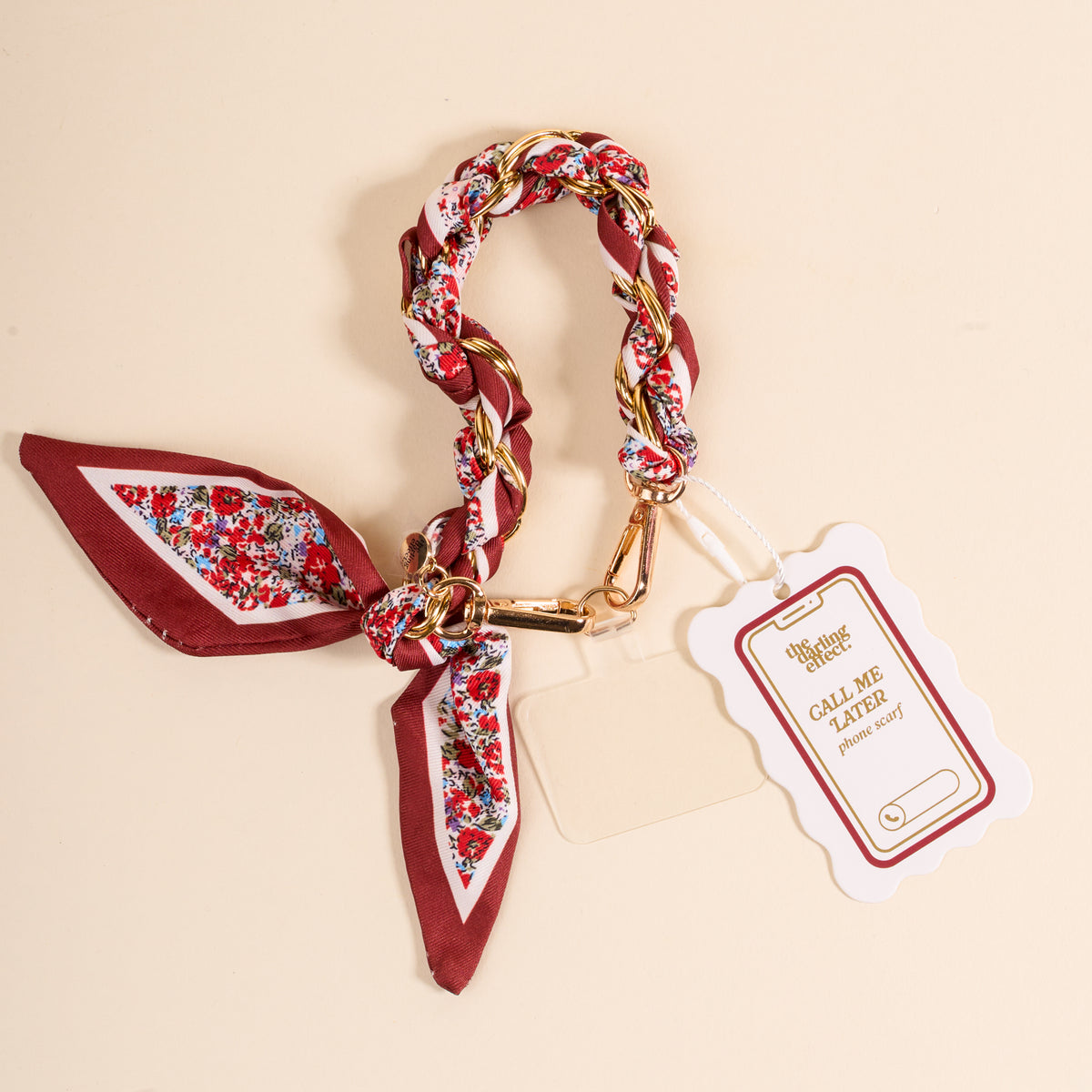 The Crimson Rose Call Me Later Phone Scarf by The Darling Effect is a braided red, white, and gold bracelet with a floral pattern, gold hardware, and tied scarf accent. It comes with a "CALL ME LATER" tag.