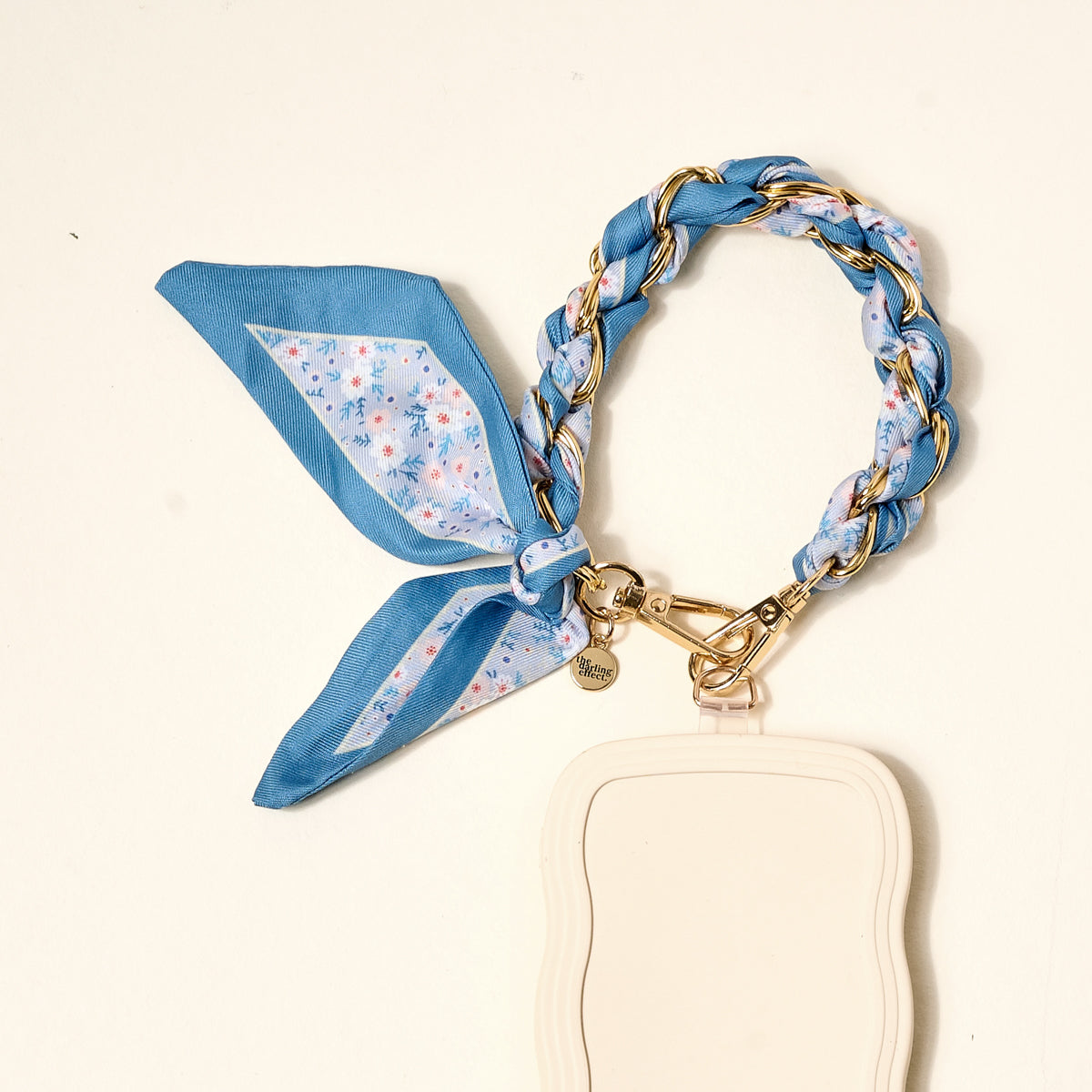 The Darling Effect's Forget Me Not Call Me Later Phone Scarf, with gold links and blue floral fabric, pairs chicly with a white scalloped-edge card holder for a stylish accessory against a light backdrop.
