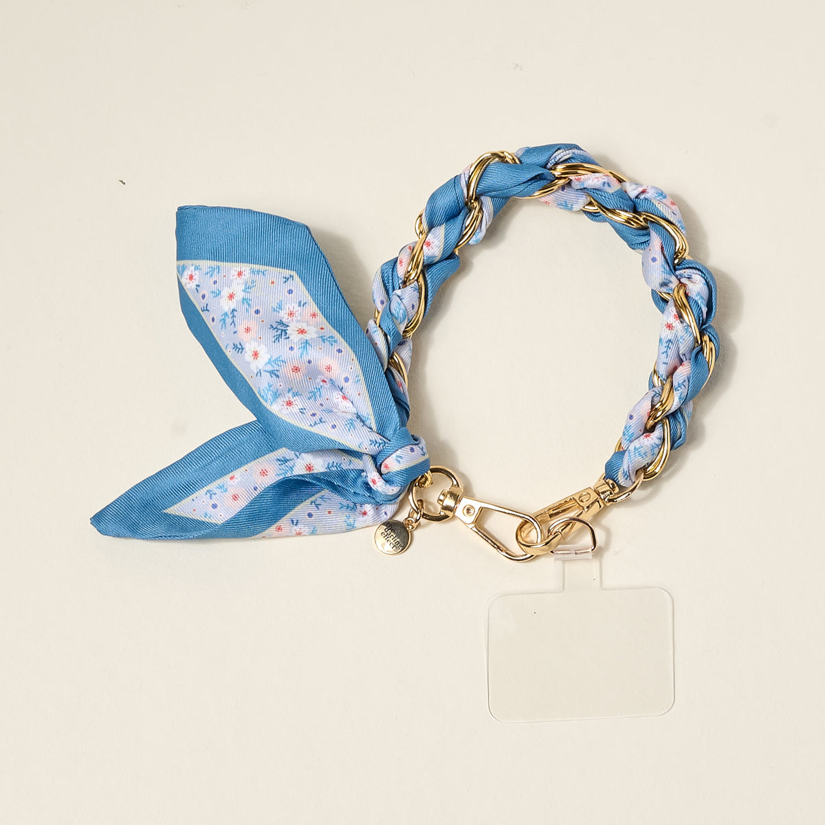 The Darling Effect's Forget Me Not Call Me Later Phone Scarf is a chic braided keychain with gold links, a blue floral scarf, metal clasp, and clear tag—perfect for adding style to your bag or securing your phone.