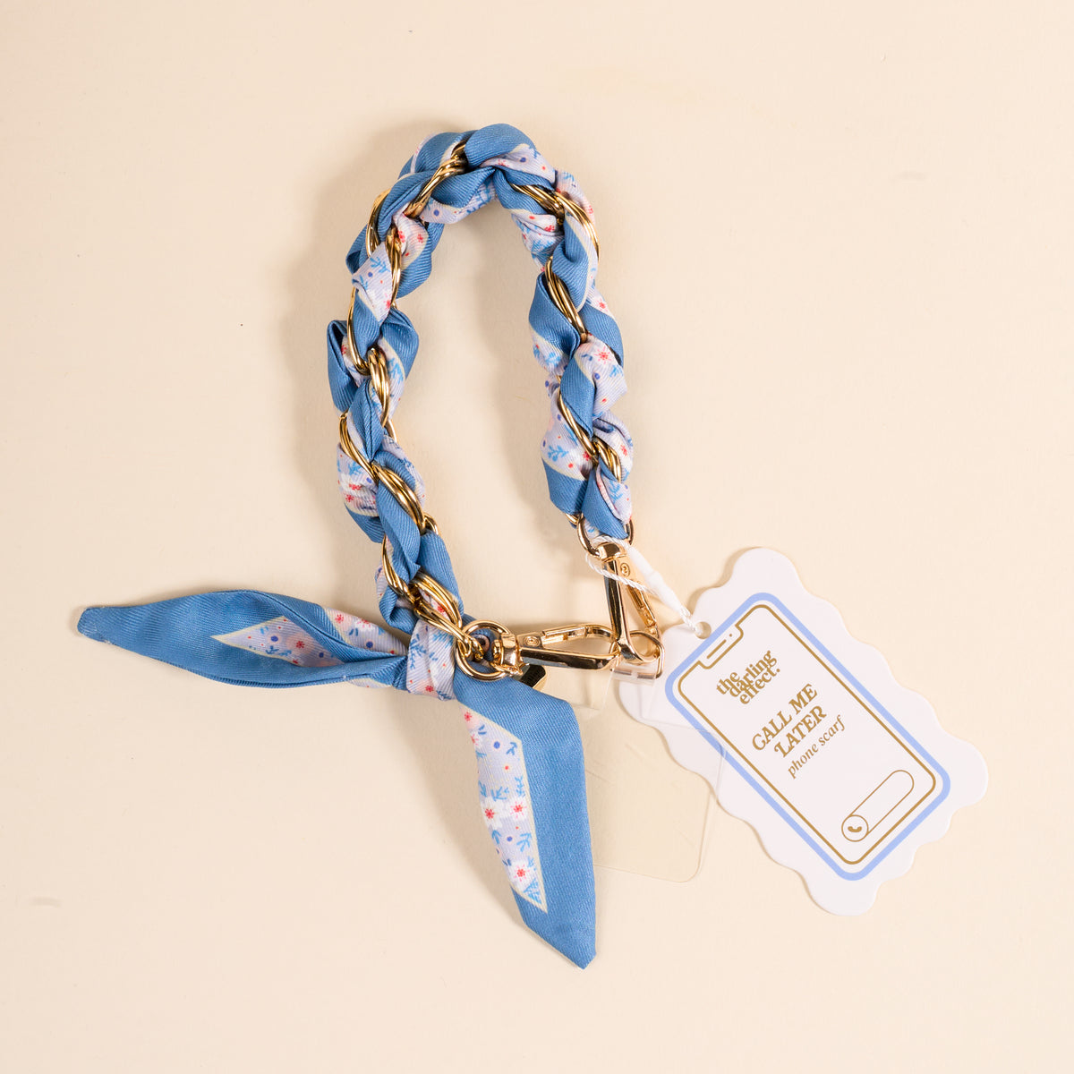 The Darling Effect’s Forget Me Not Call Me Later Phone Scarf features a blue floral braid with a gold chain, looped in a knot with secure grip and gold clasp, plus a white and gold Call Me Classy tag, displayed on a light beige surface.