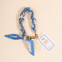The Darling Effect’s Forget Me Not Call Me Later Phone Scarf features a blue floral braid with a gold chain, looped in a knot with secure grip and gold clasp, plus a white and gold Call Me Classy tag, displayed on a light beige surface.