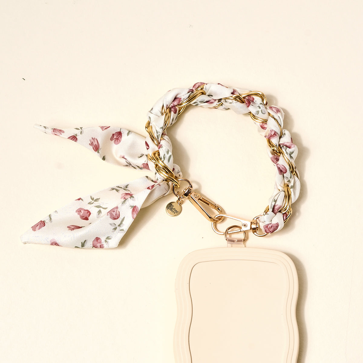 The Darling Effect’s Garden Party Call Me Later Phone Scarf features a gold chain bracelet entwined with a white floral scarf and gold clasp, attached to a beige keychain—a chic essential for your accessories.