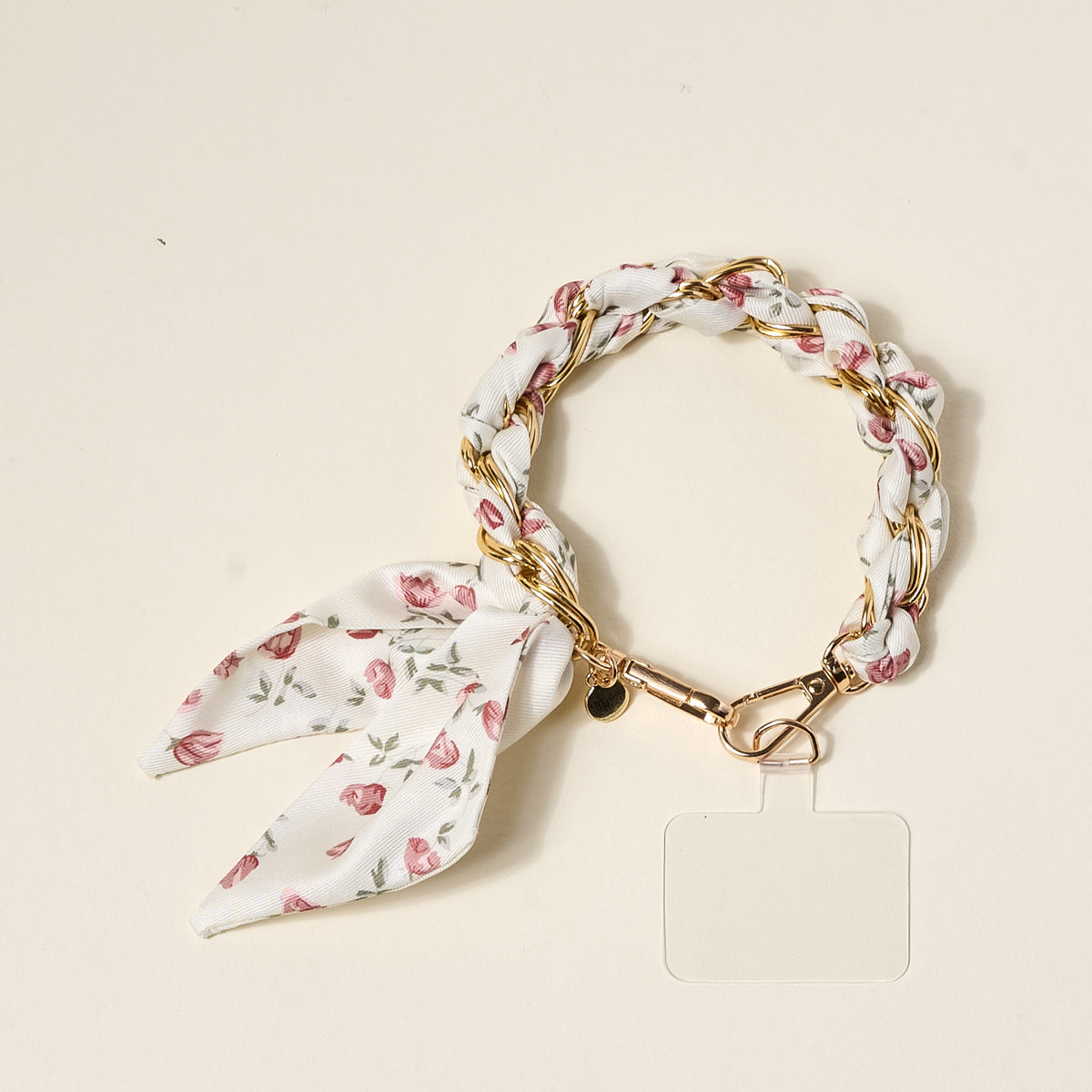 The Darling Effect's Garden Party Call Me Later Phone Scarf features a gold chain intertwined with white fabric in a pink floral pattern, finished with a bow and clear rectangular tag, displayed on a light beige surface.