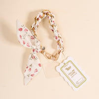 The Darling Effect’s Garden Party Call Me Later Phone Scarf is a floral keychain with a braided strap, gold clasp, and bow. On a light background, it displays a scalloped tag reading CALL ME with a phone graphic.