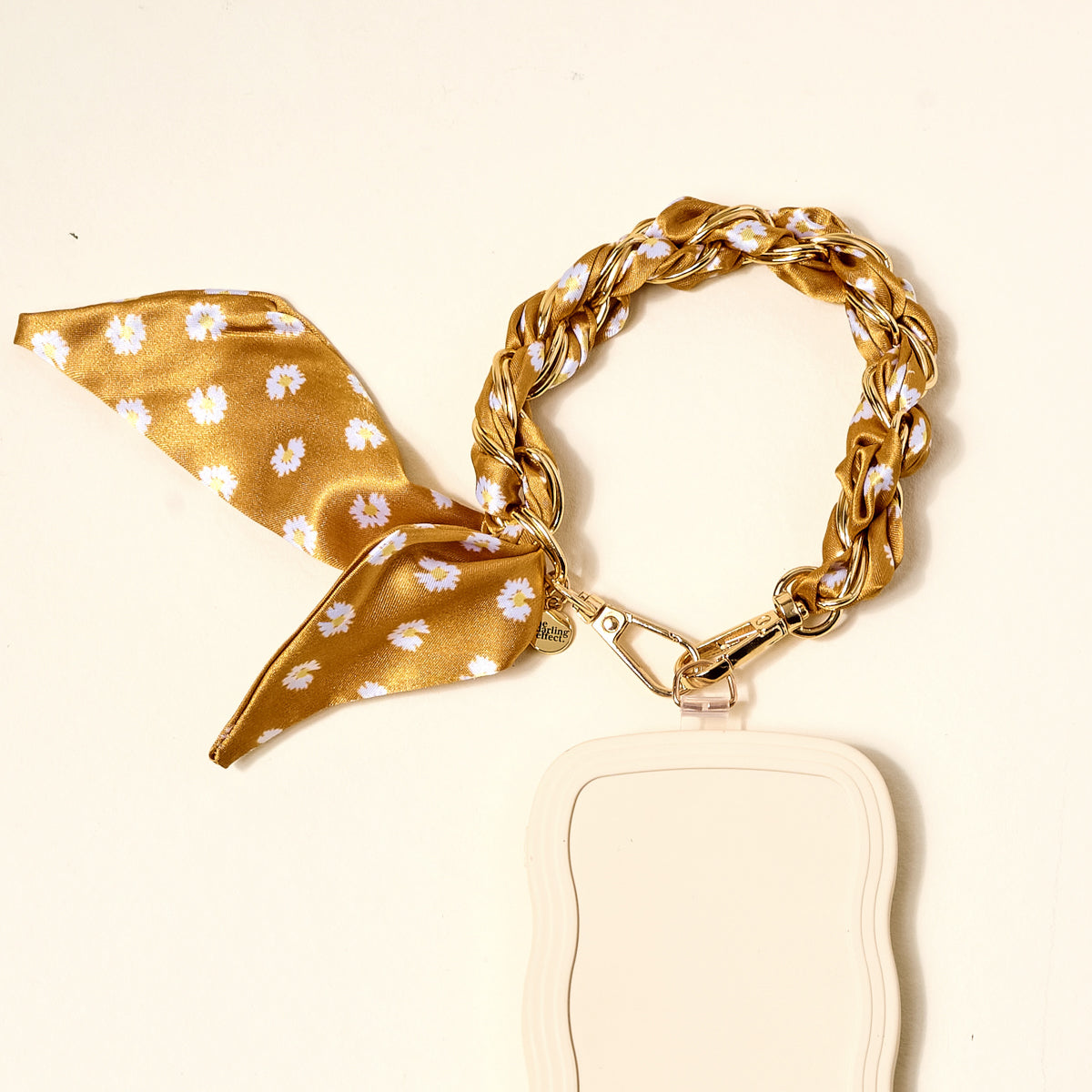 The Darling Effect's Honey Blossom Call Me Later Phone Scarf features a gold chain strap and yellow scarf with white flowers, clipped to a beige wavy-edged phone grip, displayed against a cream background.