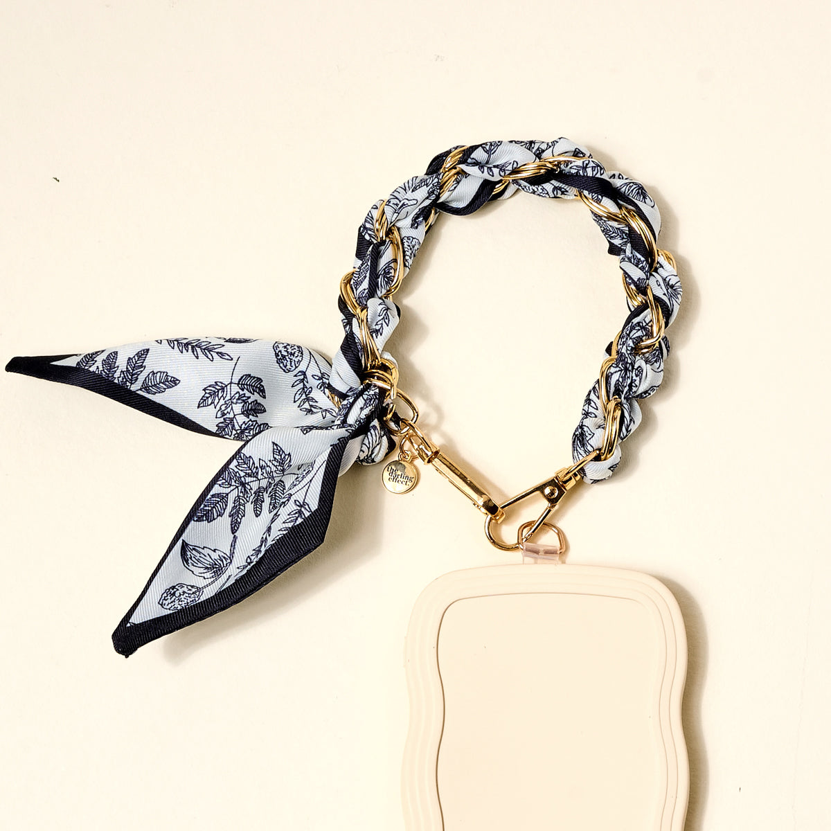 The Darling Effect Monochrome Muse Call Me Later Phone Scarf features a cream case with a gold-tone chain, black and white floral silk scarf bow, and gold clasp—an elegant accessory for your device.