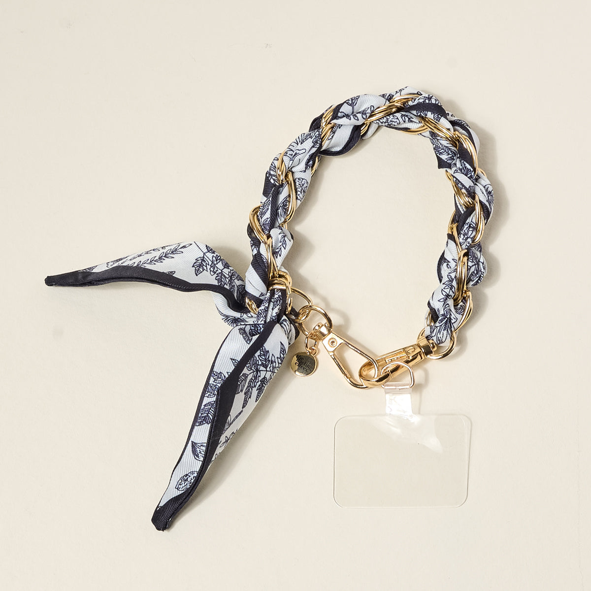 The Darling Effect's Monochrome Muse Call Me Later Phone Scarf features a gold-tone chain woven with black-and-white fabric, finished with a knot, gold lobster clasp, and a clear rectangular tag for stylish functionality.