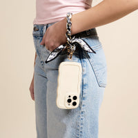 A person's hand holds a cream-colored phone with The Darling Effect's Monochrome Muse Call Me Later Phone Scarf wristlet, a stylish black and white floral accessory that doubles as a chic fashion statement.