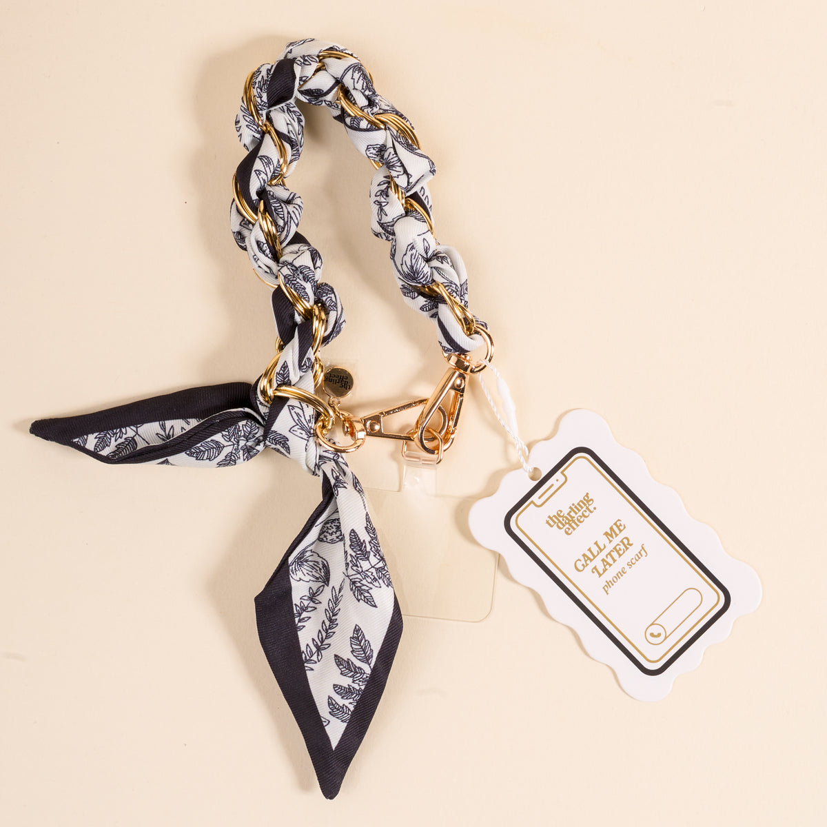 The Darling Effect’s Monochrome Muse Call Me Later Phone Scarf features black-and-white patterned fabric woven through a gold-tone chain with a bow, clasp, and “Call Me Pretty” tag—a chic accessory for any style.