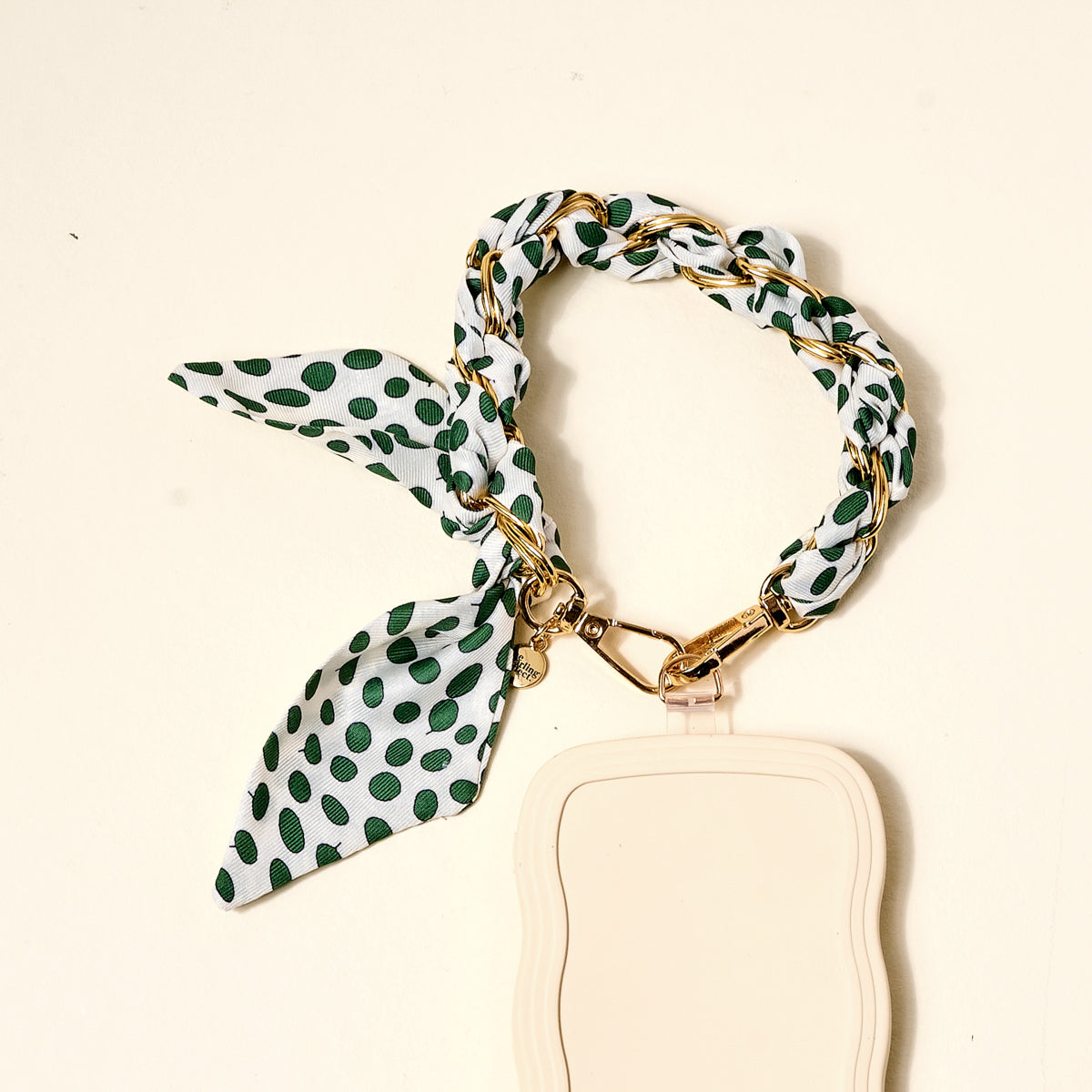 The Darling Effect's Olive Dot Call Me Later Phone Scarf features a gold-tone chain bracelet interwoven with white fabric with green polka dots, tied in a knot, and attached to a beige rectangular scarf—a chic accessory.