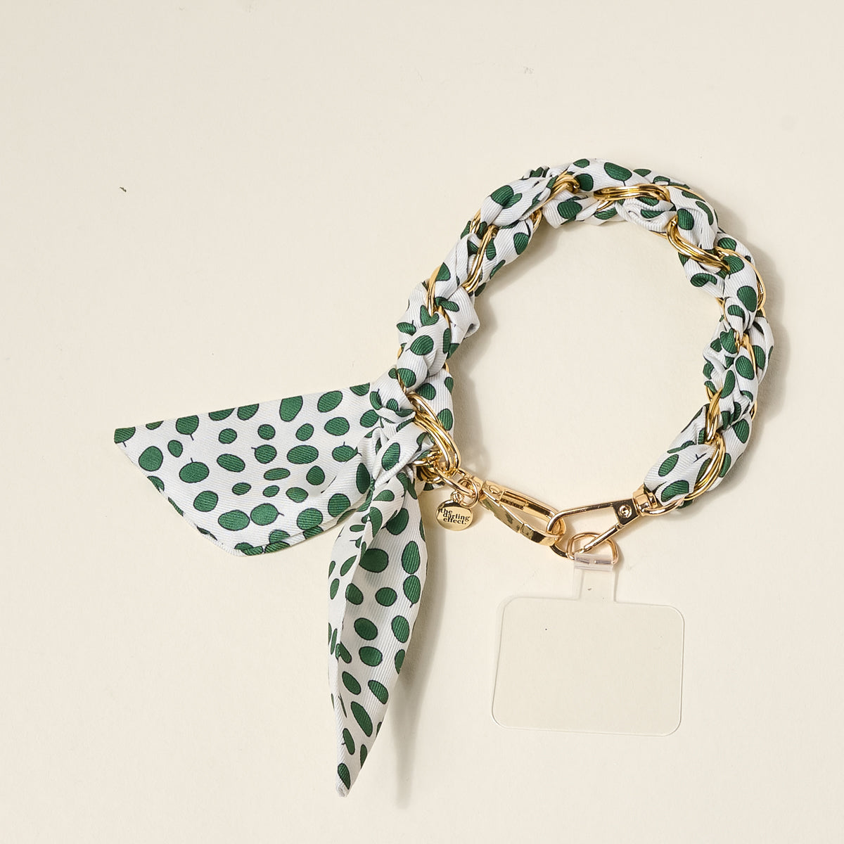 The Darling Effect's Olive Dot Call Me Later Phone Scarf features a gold-tone chain with white fabric adorned in green polka dots and a bow, accented by a small gold charm and clear tag—an elegant accessory shown on a light backdrop.