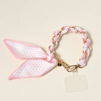 The Darling Effect’s Sugar Dot Call Me Later Phone Scarf features a gold-tone chain-link bracelet woven with pink and white polka dot fabric, a matching bow, and a clear charm—adding playful elegance to your style.